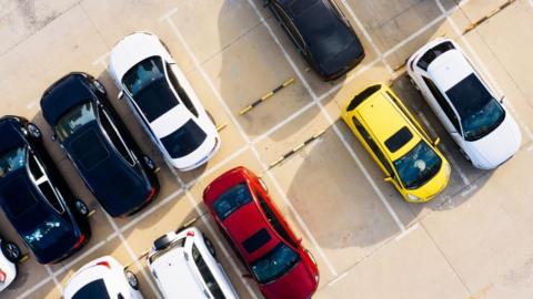 An overhead view of a car park with vehicles parked in marked bays, including black, white, red and yellow cars separated by white lines on a concrete surface.