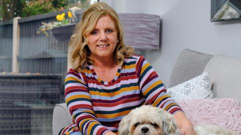 Carol Player smiles at the camera in a stripy top with a small dog. She has blonde hair to her shoulders and sits on a grey sofa in front of a window.