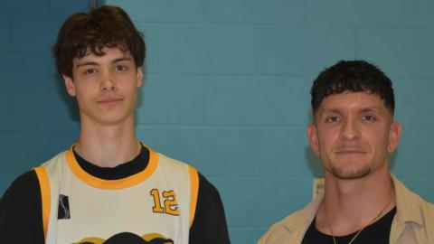 Student Isaac in a yellow and black basketball overshirt stands next to Matt Hayden who wears a beige shirt over a black T-shirt. Both are standing in front of the blue wall of the sports hall at Suffolk New College.