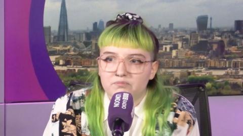 Veronique Hawksworth speaking into a purple BBC Radio London microphone with a London skyline background on the screen behind