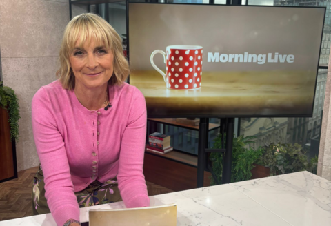 Louise Minchin leaning on the breakfast bar in the Morning Live studio holding cue cards.