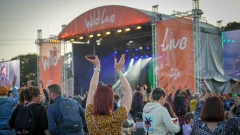 A woman in a yellow top with red hair and sunglasses on her head stands in a large crowd in front of the main stage at music festival 'Wild Live' at Yorkshire Wildlife Park in Doncaster
