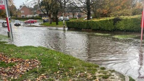 A residential street that has flood water completely covering it. There is a bus stop sign on the left, and grass next to it. There is a black car driving towards the water in the distance