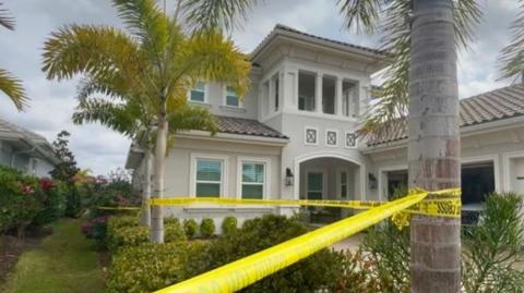 A cream Florida home, with a number of palm trees in the foreground. There is yellow police tape wrapped around the trees. In the garden in front of the house there are a range of bushes, and a patio car parking space.