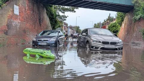 A blue Ford car and a silver BMW car appear to be motionless in floodwater below a railway bridge. In the foreground is a green crocodile inflatable. In the background are some people and houses.