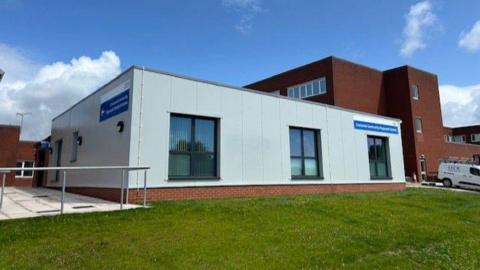 The Croslands Community Diagnostic Centre. It is a square grey building with a blue sign which states its name. Furness General Hospital can be seen behind it and a green lawn stands in front of it.