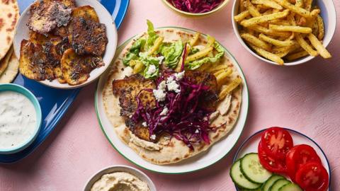 A plate with flatbread, grilled tofu, chips and salad on a pink background with a plate of tofu, salads and bowl of chips.