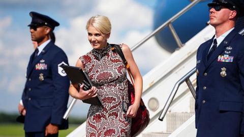 Natalie Harp walks off Air Force One wearing a sleeveless printed red dress. She is smiling and has a laptop in her hands.