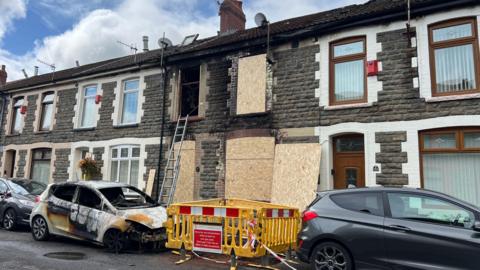 A stone terraced house with fire damage around the windows and doors, and wooden boards covering them. In front are two cars parked, one is badly fire damaged.