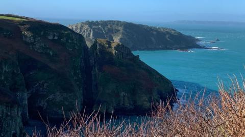 Two grass covered rocky headlands project out to the sea from the left of the shot. It is a sunny day and the sun is shining. 