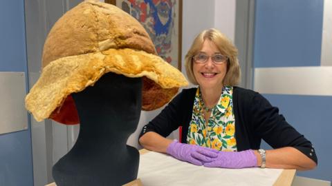 Conservator Jacqui Hyman, who is wearing a floral blouse and purple protective gloves, smiles as she sits alongside the newly conserved hat. The hat is made of ochre-coloured felt, with a wide brim. 