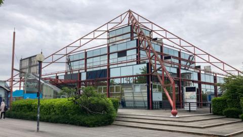 A glass building shaped like a pyramid with red metal support beams lining the edges, this is The Point in Milton Keynes.