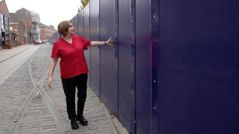 A woman wearing a bright red short-sleeved shirt and black trousers is standing on a cobblestone street next to a tall, solid barrier made of dark blue panels. She is gesturing toward the barrier with one arm extended, as if indicating its length or height. The street has old metal tram lines running along its surface, and older brick buildings are visible in the background on the left side.
