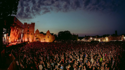 A large crowd of people in the dark with a castle illuminated in the background and a stage on the left hand side