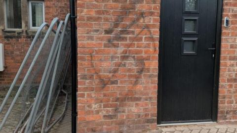 A swastika is painted in black onto a brick wall with a black door alongside it. There are metal fence panels to the side, leaning against the wall.