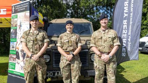 Three army personnel stand in front of a military Land Rover in khaki uniform. The two men and a woman are all wearing black berets and standing with their hands clasped in front of them, with banners either side promoting Army Reserves.
