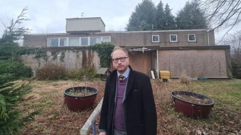 A man in a black coat, purple jumper, blue shirt and orange tie is standing outside a boarded up building. The building is covered in ivy, the grass and plants outside are dying and water is running down the side of one of the walls.