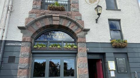 A historic pub frontage with a stone archway entrance and a sign reading “Thomas De La Rue” above the door. The building has an off white plaster walls with grey stone accents, window boxes filled with green plants and yellow flowers, and a street sign for “North Plantation” on the left. The red entrance door is open above which hangs a large wall lamp. A glass winder reflects trees and sky outside.