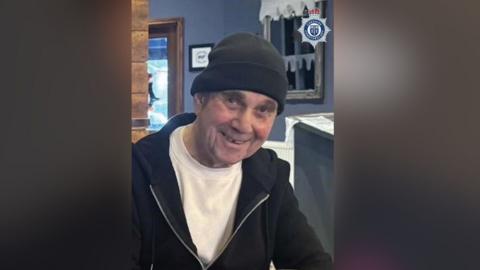 John McColl is seen smiling waring a black beanie hat, t-shirt and black hoodie.