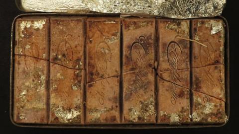A 126-year-old Cadbury chocolate bar that sits within a tin of the same rectangular shape. The bar has been cut into six rectangular sections, some of which have broken over time. The Cadbury logo is embossed on each chocolate rectangle. Some foil is stuck on the chocolate, which has a variety of brown hues.
