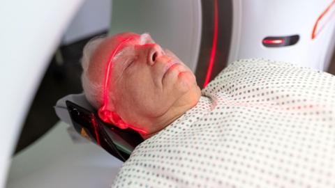 An older man lies down wearing a white hospital gown as he undergoes radiation treatment. There is a large red laser line across his forehead and another line going down his nose.