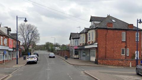 A crossroads on a main road, with shops on either side of the road, and the hump of a railway bridge in the distance