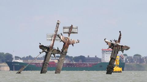 Three masts covered in bird droppings stick out of the sea. Behind is a large tanker and a yellow buoy