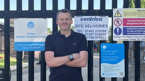 A man in a dark polo shirt stood in front of a large metal gate. He is crossing his arms. The sign on the gate is for Horley Sewage Treatment Works and has the Thames Water logo on it.