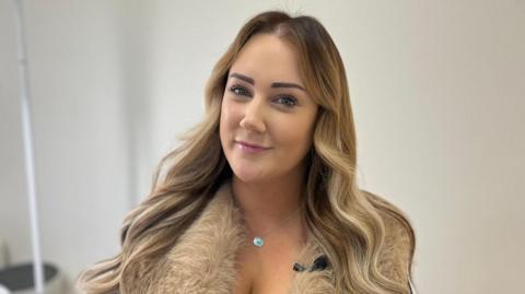 Lizzie has long, wavy, highlighted brown hair. She is wearing a faux fur gilet and black top, with a necklace and is smiling at the camera. She is sat in a white clinical treatment room. It is a head and shoulders shot of her.