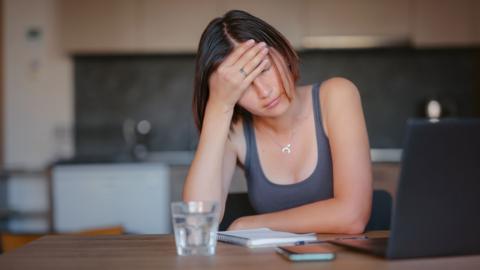 Young woman with her head in her hand, sitting at a desk with a laptop, feeling overwhelmed - stock image