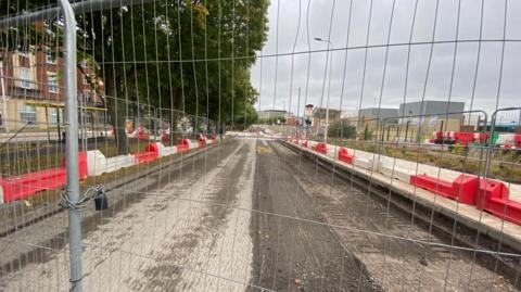 A road fenced off behind metal fencing. The road surface is under construction and is lined by red and white plastic barriers.