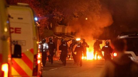 A large group of police officer dressed in riot gear and holding sheilds walking towards a fire and smoke outside a row of houses. Two police vans can be seen behind them.