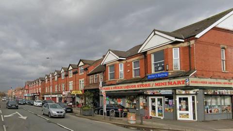 Princess Road, Moss Side, showing a row of shops including Richie & Nickie Liquor Store/Mini Mart and some cars travelling on the road on a dry day with grey clouds.