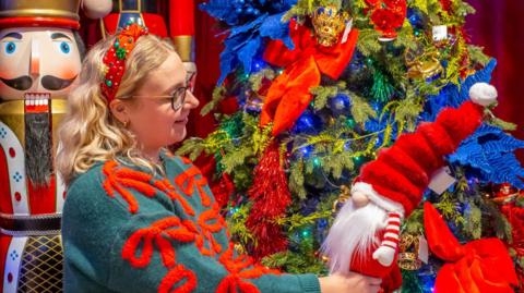 A woman with blonde hair wearing dark framed glasses and a green and red Christmas jumper is stood in front of a Christmas tree holding a toy father Christmas.