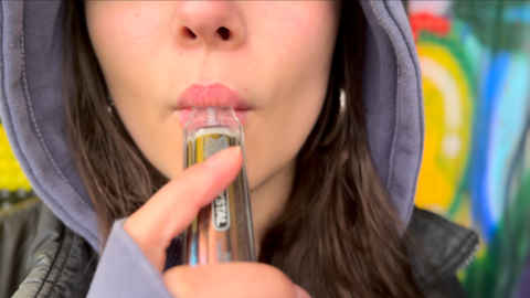 An anonymous teenage girl holds a vape to her lips, just the lower half of her face visible. She is in a purple hoodie, hood up, and black jacket and has long brown hair. She is in a park with colourful graffiti behind her.