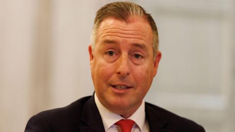 A man with grey and black hair, he is wearing a dark blazer, white shirt and red tie