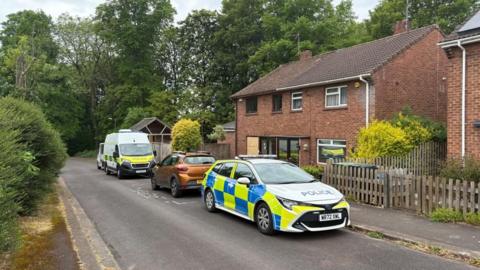 Two police vehicles are parked outside a red-brick, two-story house at the end of a quiet residential street.