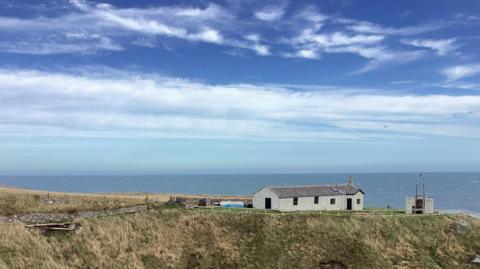 Remote buildings sit on a coast beneath a partly cloudy sky