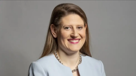 Head-and-shoulders studio portrait of a woman against a plain light grey background. The person has long brown hair parted to one side, visible teeth in a broad smile, and is looking toward the camera. The person is wearing a light blue jacket or blazer-style top and a chunky silver chain necklace. Soft, even lighting creates a professional corporate headshot appearance.
