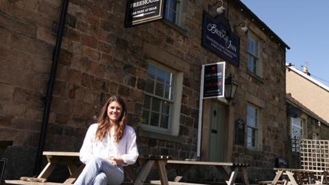 Elisa Hargreaves sitting on a bench in front of the Bridge Inn which is a stone building with a "for sale" sign in front of the door. Elisa has long brown hair and is smiling into the camera with her hands in her lap. She is wearing a white shirt and light blue jeans.