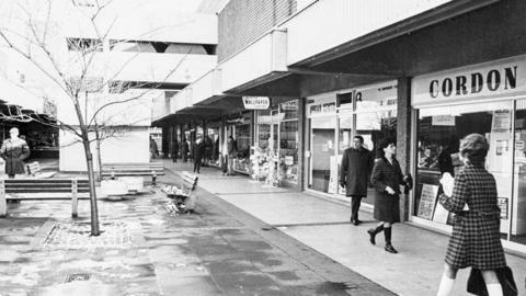 A black and white picture of a shopping centre with people walking around. There is a tree in the middle of a walkway and benches near it. There is a shop on the right called Cordon.