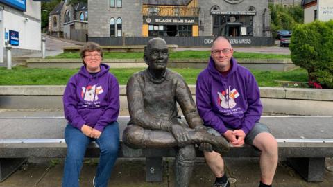 Roz and Lee Harris sitting on a bench at Fort William. They are sitting beside the statue "Sore Feet" which depicts a walker rubbing his feet. There are buildings in the background.