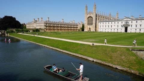 An aerial shot of a man punting a couple along the River Cam and past King's College and a large white building. There are a couple of people walking along a path in front of the buildings.