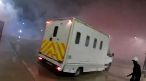 A screenshot of the police van seen on the Elbit Systems UK site. The van is a large white vehicle with small windows and a chevron pattern across the back. A figure wearing black with a white helmet can be seen in the lower right corner of the image holding a long unidentified object. 