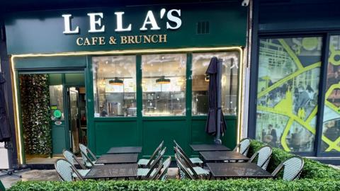 Cafe tables and chairs outside a green, gold and white cafe front. They are surrounded by a green hedge.