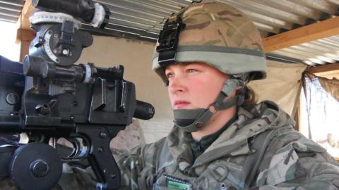 Nadine Askew wearing a camouflage military uniform and rounded helmet with a chin strap. She is sitting underneath a corrugated metal shelter, looking towards the left as she aims a machine gun.
