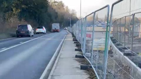 The A134 with metal fencing to the right, showing a housing estate being built, and cars driving along the road on the left. There are about four cars on the road, including one lorry. 