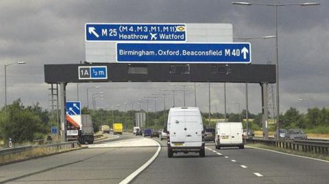 Vehicles in two lanes on the M40 travel under a large blue sign that directs them either to Heathrow and Watford or Birmingham, Oxford and Beaconsfield. The picture has been taken on a stormy day.