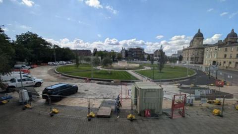 Construction work taking place at Queens Gardens, with lawns and a fountain in the background and construction vehicles and cabins in the foreground.