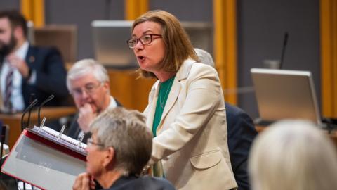 Eluned Morgan stood in the Senedd debating chamber wearing a cream jacket and a green top, while her ministerial colleagues are sat around her. 